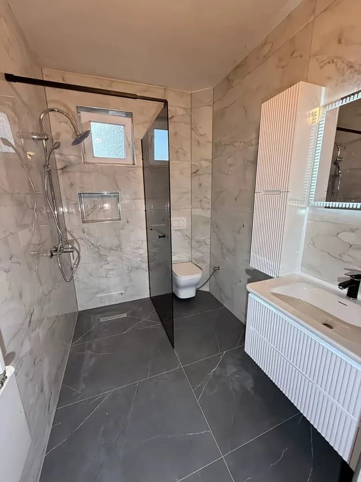 Teslim edilen banyo: beyaz mermer görünümlü duvarlar, antrasit zemin, cam duş bölmesi ve beyaz lake dolap